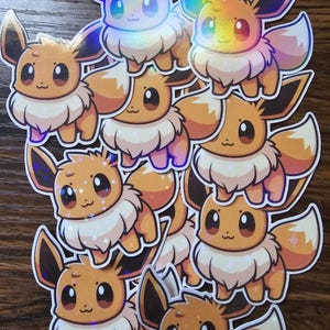 May include: A set of ten holographic stickers featuring a cartoon Eevee with a brown and orange colour scheme. The stickers are glossy and have a sparkly finish.