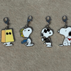 May include: Four Snoopy character charms with silver clasps. The charms depict Snoopy in different outfits, including a yellow block, a blue scarf, sunglasses and a "Joe Cool" shirt, and holding a heart. The charms are white, black, yellow, and blue.