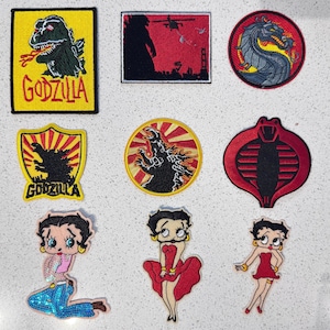 May include: A collection of nine embroidered patches featuring Godzilla imagery, a dragon emblem, a cobra logo, and three distinct depictions of Betty Boop. The patches vary in shape, including rectangular, circular, shield, and character-based outlines.