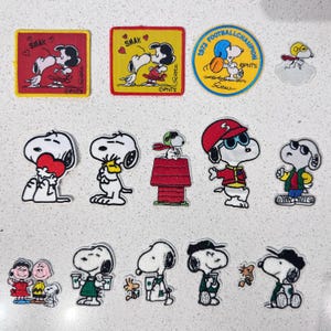 May include: A collection of fifteen embroidered Peanuts character patches featuring Snoopy and friends in various poses, including square, circular, and die-cut shapes with vibrant colours like red, yellow, and blue, showcasing iconic cartoon scenes.