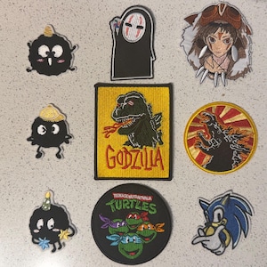 May include: A selection of embroidered patches showcasing pop culture icons. Includes a Godzilla patch with the word "GODZILLA", a Teenage Mutant Ninja Turtles patch, and a Sonic the Hedgehog patch.