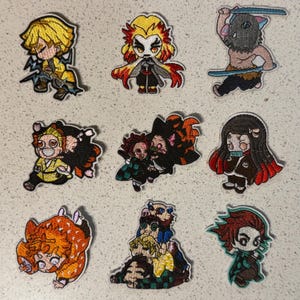May include: Nine embroidered patches featuring anime characters. The patches showcase characters with vibrant colors, including yellow, red, and black. Each patch has a unique design, with some holding weapons or in action poses. The patches are on a light-colored surface.