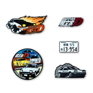 May include: A set of five enamel pins featuring Initial D themes: a black and white car with orange flames, an Initial D logo, a rectangular Japanese license plate with text, a circular badge with three cars, and a drifting black and white car.