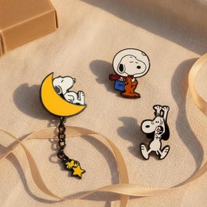 May include: Three Snoopy enamel pins. One pin shows Snoopy sleeping on a yellow crescent moon with a dangling star. Another shows Snoopy in an astronaut helmet and suit. The third pin depicts Snoopy with arms raised.