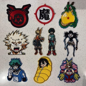 Könnte beinhalten: Eine Sammlung von neun bestickten Aufnähern mit verschiedenen Anime-Charakteren und Symbolen, darunter ein roter Ouroboros, ein weißer Kreis mit dem schwarzen Kanji für Magie, ein grüner Drache mit gelber Kugel und diverse Figuren.