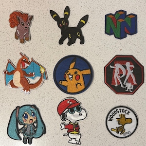May include: Assortment of embroidered patches featuring cartoon characters and logos. Includes a brown and orange fox-like creature, a black cat-like creature, a green, red, and blue N64 logo, a red and orange dragon, a Pikachu patch, and more.