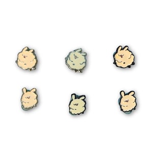 Puede incluir: Un conjunto de seis pequeños pines de esmalte con cabezas de conejo de dibujos animados en varios tonos crema y amarillo pálido. Cada pin muestra una expresión facial diferente con detalles de líneas negras para ojos y bocas.