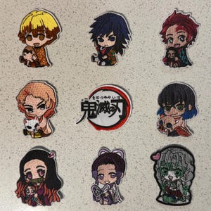 Könnte beinhalten: Neun aufgestickte Aufnäher mit Anime-Charakteren. Die Aufnäher zeigen verschiedene Charaktere mit detaillierten Gesichtsausdrücken und farbenfrohen Outfits. Der zentrale Aufnäher zeigt japanischen Text in einem roten und weißen kreisförmigen Design. Die Aufnäher sind auf einer hellen Oberfläche angeordnet.