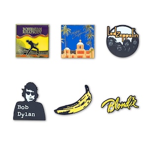 May include: A collection of six enamel pins featuring music-themed designs: a square Bohemian Rhapsody pin, a square Hotel California pin, a circular Led Zeppelin pin, a Bob Dylan portrait pin, a yellow banana pin, and a yellow Blondie logo pin.