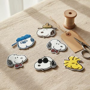 May include: A collection of embroidered Snoopy patches in various designs, including Snoopy with a blue cap, sunglasses, and a red bow tie. A wooden spool of thread, scissors, and a tag are also present.