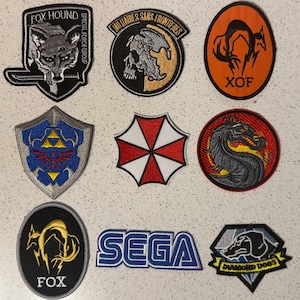 Puede incluir: Una colección de parches bordados con varios diseños. Se incluyen parches con un zorro, una calavera, un dragón, un escudo, un paraguas y el texto "SEGA". Los parches son de varios colores, incluyendo negro, naranja, rojo, azul y blanco.