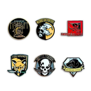 Puede incluir: Colección de seis pines esmaltados con varios diseños. Los pines incluyen una calavera, un zorro y un perro, junto con textos como "DEATH STRANDING" y "DIAMOND DOGS". Los pines son de varias formas y colores.