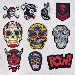 May include: A collection of ten embroidered patches featuring a skull with crossbones, a dog, a cat, a skull emblem, three sugar skulls with floral and geometric patterns, a red horned mask, and a black patch with the red text POW!