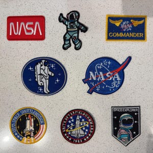 May include: A collection of seven embroidered patches featuring space themes. Included are a red NASA logo, a blue astronaut, a blue NASA Commander patch, a blue oval astronaut patch, a blue NASA meatball logo, a space shuttle patch, and a shield.
