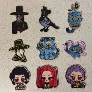 May include: A collection of embroidered patches featuring various designs. The patches include a figure in a black hat, a crow, several blue cat designs, and cartoon figures with large eyes. The patches are in various colors, including black, blue, yellow, and pink.