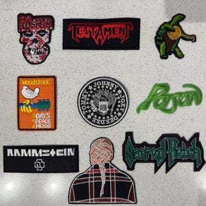 Könnte beinhalten: Eine Sammlung von acht gestickten Aufnähern mit verschiedenen Motiven, darunter ein Misfits-Totenkopf, ein Testament-Logo, eine Banjo spielende Schildkröte, eine Woodstock-Grafik, ein Ramones-Siegel, ein Poison-Logo, ein Rammstein-Logo und eine Person mit Zopf.
