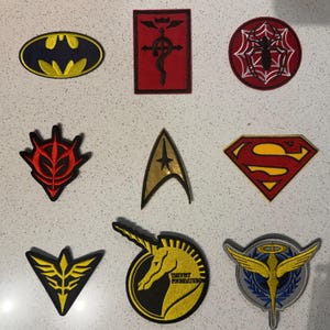 May include: A collection of embroidered patches featuring various pop culture and fictional symbols. Includes a Batman logo, a Star Trek insignia, a Superman logo, and other emblems in various colors and shapes.