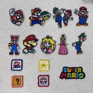 May include: A collection of embroidered fabric patches featuring Super Mario characters including Mario, Luigi, and Princess Peach, along with pixelated icons like a star, mushroom, question block, and the Super Mario logo text.