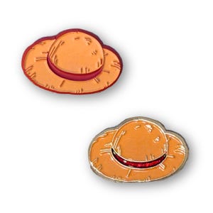 May include: Two orange enamel pins shaped like a straw hat, each with a red band. The pins are set against a white backdrop.