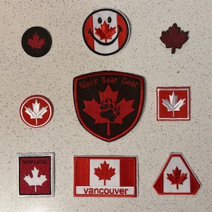 Peut inclure: Collection de patchs brodés avec des symboles canadiens. Les motifs comprennent des feuilles d'érable, le drapeau canadien et du texte tel que "Black Bear Gear" et "Vancouver". Les patchs sont rouges, blancs et noirs.