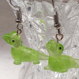 May include: A pair of lime green lizard earrings. Each earring features a small, translucent lizard charm with a detailed design. The earrings hang from silver-colored fish hook ear wires. The earrings are displayed in a clear glass vase.