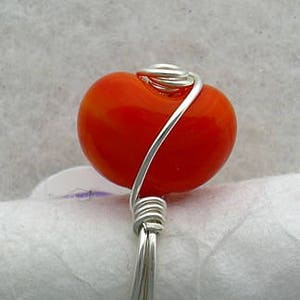 May include: A silver wire ring with an orange glass bead. The bead is wrapped in wire and has a swirl design.