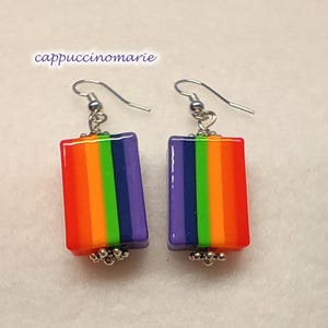 May include: A pair of drop earrings featuring rectangular beads with vertical stripes in purple, dark blue, green, orange, and red. The beads are accented with small silver-toned metal beads at the bottom and silver-toned ear wires. Text reads cappuccinomarie.