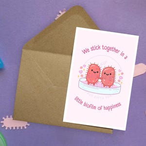 May include: A greeting card with the text "We stick together in a little biofilm of happiness" featuring two smiling pink bacteria. The card is inside a brown envelope, surrounded by pink microbe cutouts and laboratory tools on a purple surface.