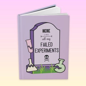 May include: A small, open book with a lilac cover. The cover features a tombstone design with the text "HERE lies all my FAILED EXPERIMENTS" and illustrations of beakers and a skull and crossbones.