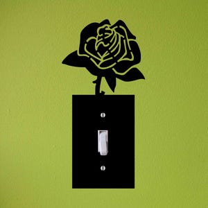 May include: A black rose-shaped wall decal above a black light switch cover. The light switch is white, and the wall is painted a light green color. The rose is a silhouette with detailed cutouts.