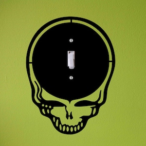 May include: Black skull-shaped light switch cover with a white switch and a lime green wall background. The skull design has a circular head and a detailed jawline. The switch is in the center of the skull's head.