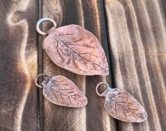 May include: Three copper leaf pendants with etched details and a loop for hanging. The leaves vary in size, with the largest measuring approximately 2.5 inches long. The pendants are displayed on a rustic wooden surface.