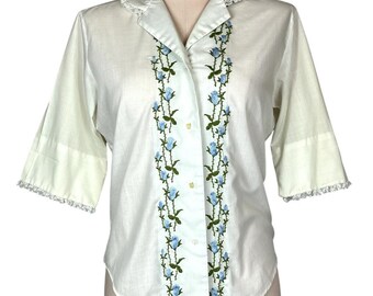 Puede incluir: Una camisa blanca de botones con un ribete de encaje en el cuello y las mangas. La camisa tiene un diseño de bordado floral con flores azules y tallos verdes.