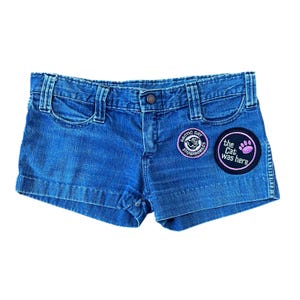 Peut inclure: Short en jean bleu avec deux patchs. Un patch est un cercle noir avec du texte blanc qui dit "the cat was here" et une empreinte de patte rose. L'autre patch est un cercle noir avec du texte blanc qui dit "Arctic Cat Snowmobiles" et une tête de chat noir et blanc.