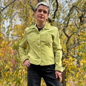 Peut inclure: Une personne portant une veste en daim vert lime à boutons et à manches longues. La veste a deux poches à rabat sur la poitrine. La personne est debout devant un arbre aux feuilles jaunes.