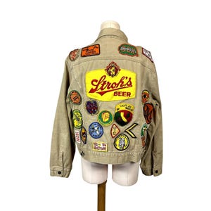 May include: A tan denim jacket with a variety of colorful patches sewn on the back. The largest patch is yellow with red and black lettering that reads "Strohs's Beer".