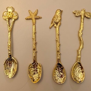 May include: Four decorative gold-colored spoons with nature-inspired designs. One spoon features a flower, another a starfish, a third a bird, and the last a dragonfly. Each spoon has a detailed handle and bowl, perfect for serving or display.