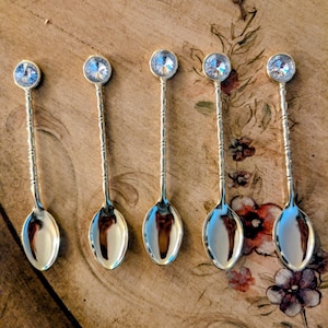 May include: Five silver-colored spoons with a decorative design on the handles and a large, clear crystal at the top of each spoon. The spoons are arranged on a wooden surface with floral accents.