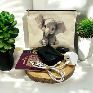 May include: A beige canvas pouch with a cute grey elephant illustration. A black wallet, passport, and white charging cable are on a wooden surface. A white charger and small green plants are also present.