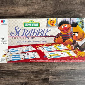 Pode incluir: Caixa do jogo de palavras cruzadas Scrabble da Vila Sésamo. A caixa apresenta o logotipo da Vila Sésamo, o Monstro das Bolachas e o Óscar. O jogo é para crianças dos 3 aos 5 anos e inclui quatro tabuleiros de jogo de dupla face. O jogo foi concebido para 1 a 4 jogadores.