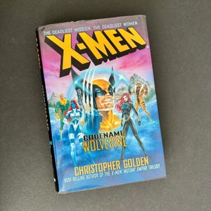 Puede incluir: Un libro de bolsillo titulado "X-Men: Codename Wolverine" de Christopher Golden. La portada presenta ilustraciones de varios personajes de X-Men, incluyendo a Wolverine, Mística y otros, con una vibrante combinación de colores amarillo, azul y negro.