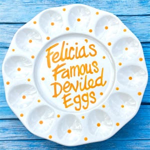 May include: White ceramic deviled egg serving platter with 12 egg indentations. The platter has orange polka dots around the edge and the words "Felicia's Famous Deviled Eggs" in orange script in the center.