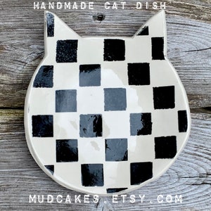 May include: Handmade ceramic cat dish with a black and white checkered pattern. The dish is shaped like a cat's head with large ears.