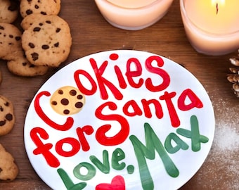 May include: A white ceramic plate with the words "Cookies for Santa Love, Max" in red and green lettering, with a cookie illustration. Chocolate chip cookies and lit candles are in the background.