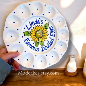 May include: A white ceramic deviled egg serving platter with a scalloped edge and blue polka dots. The center features a hand-painted yellow sunflower with green leaves and the blue text "Linda's Famous Deviled Eggs" written in a script font.