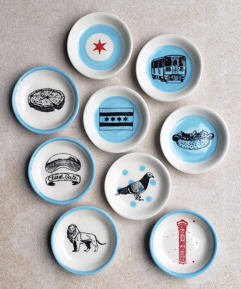 May include: Set of ten small blue and white ceramic dishes with various designs, including a pizza slice, a hot dog, a train, a pigeon, a lion, a star, and the words "Cloud Gate" and "Chicago".