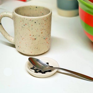 May include: A white speckled ceramic mug with a handle and a silver spoon on a white ceramic saucer with a black design.
