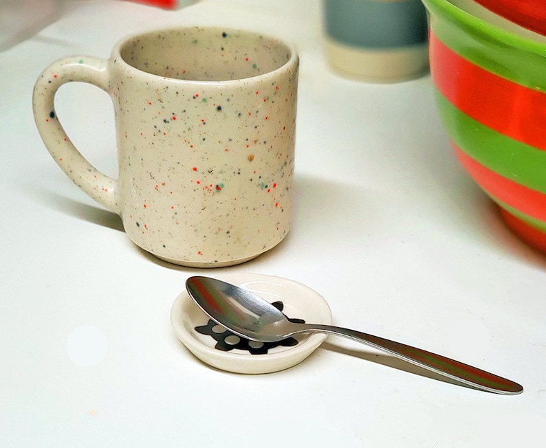 May include: A white speckled ceramic mug with a handle and a silver spoon on a white ceramic saucer with a black design.