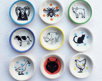 May include: Nine small, round ceramic dishes arranged in a grid. Each dish features a unique black ink illustration of an animal, including a ram, bee, pig, cow, rhinoceros, cat, goose, cat face, and chicken, with some accented by colored rims or dots.