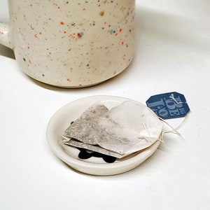 May include: A white ceramic teacup with a speckled pattern and a white ceramic saucer with a black flower design. A used tea bag sits on the saucer with a blue tea tag that reads "16 Tea for 1".